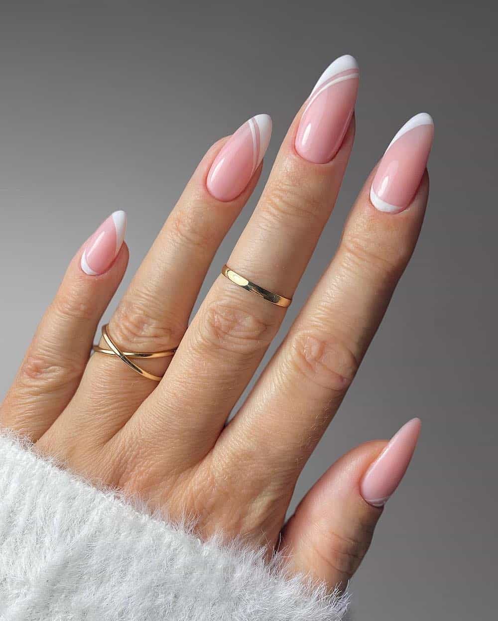 A hand with medium-length almond nails painted a pastel pink with asymmetrical white French tips and line accents