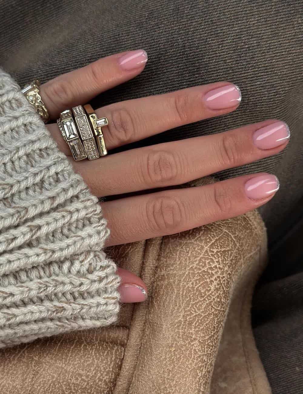 A hand with short squoval nails painted a nude pink with thin silver glitter French tips