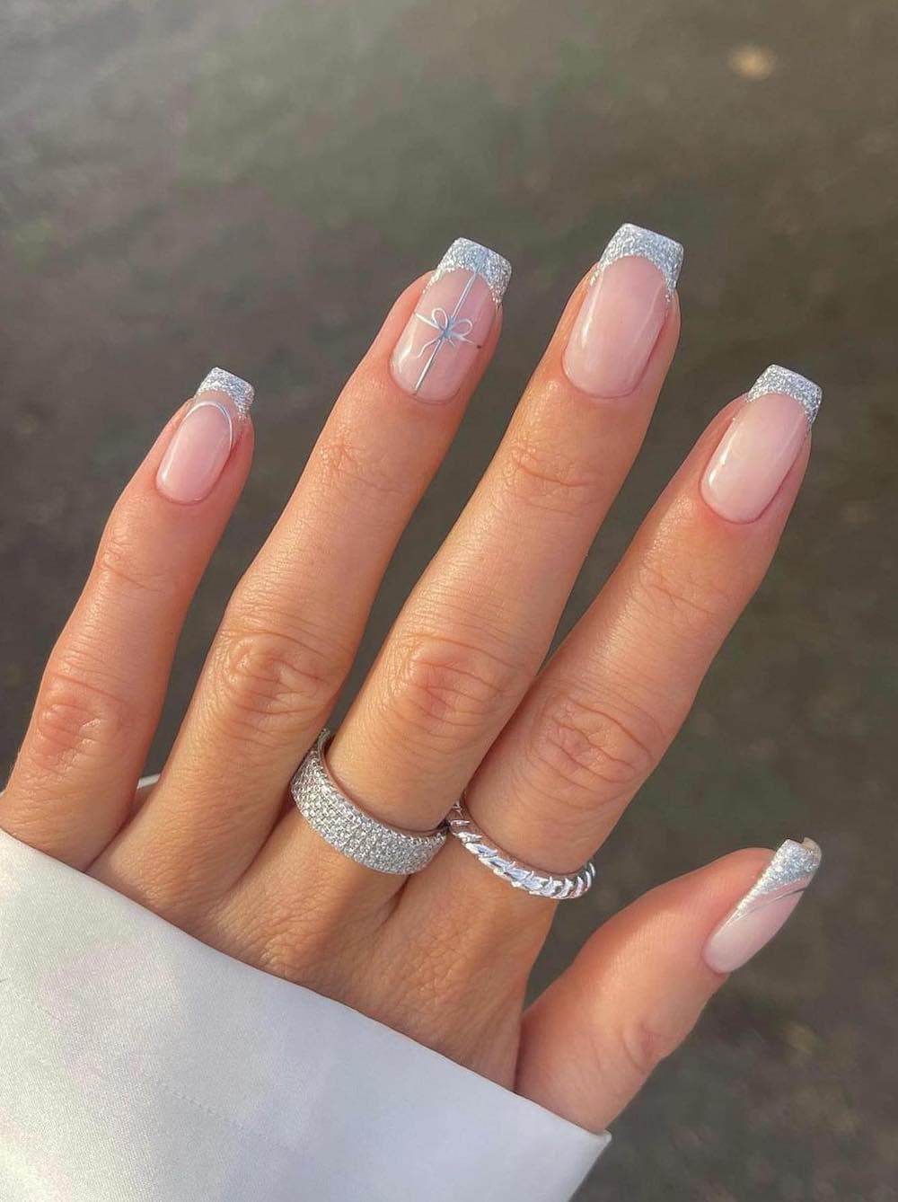 A hand with short square nails painted a glossy nude with silver glitter French tips and a bow accent nail