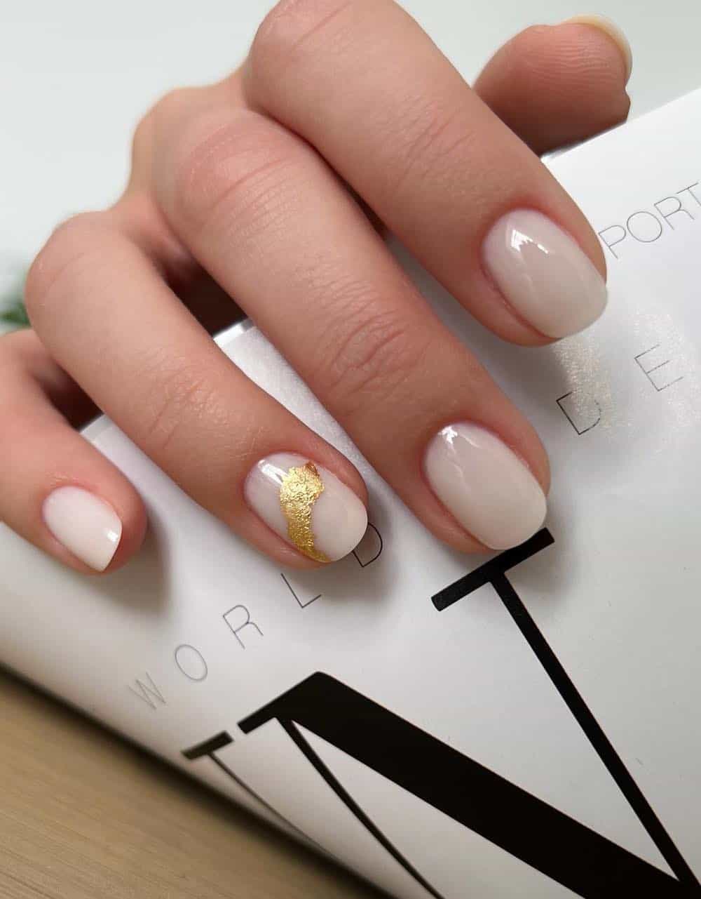 A hand with short round milky white nails with a gold foil accent nail