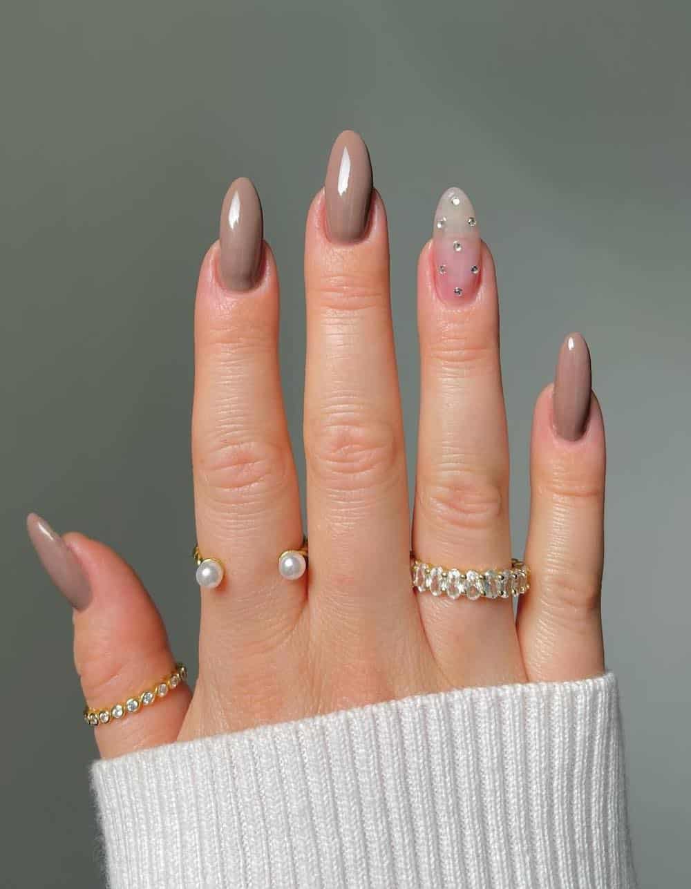 A hand with long almond nails painted a milk chocolate brown with a nude accent nail featuring gem details