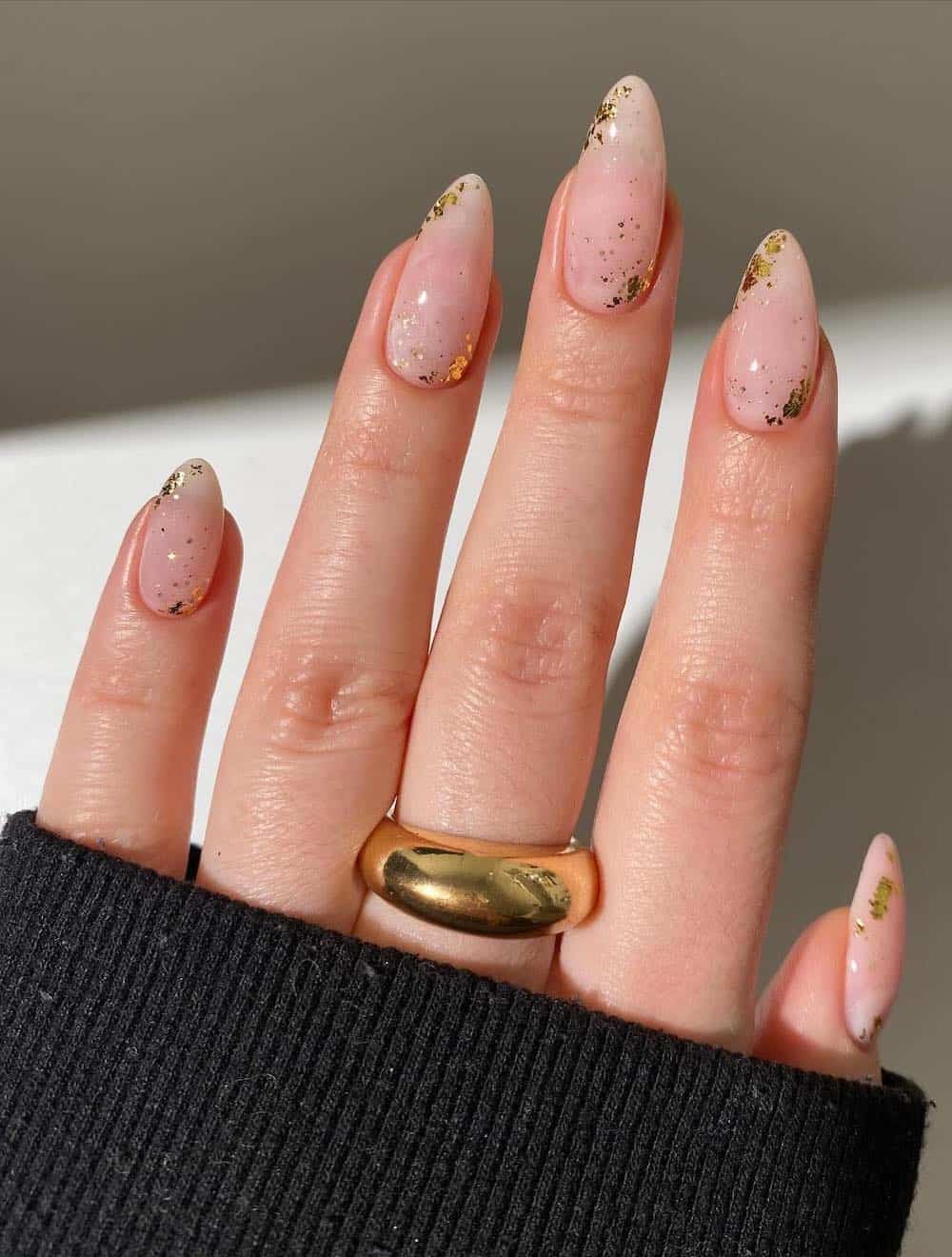 A hand with medium nude almond nails with gold flake details