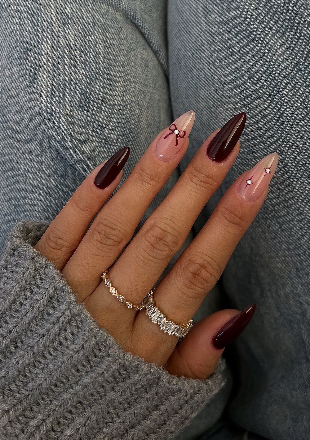 A hand with long almond nails painted a deep red with nude accent nails featuring sparkles, bows, and crystals