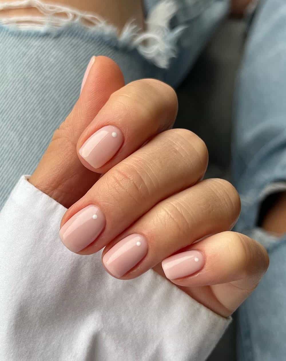 A hand with short square nails painted a nude color with a single white dot near the base