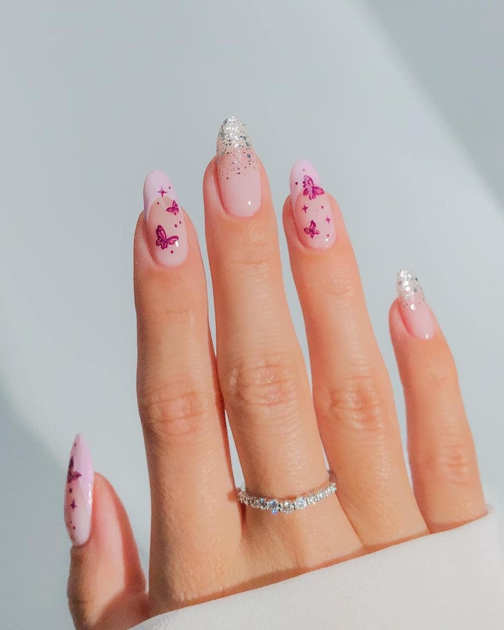 A hand with long almond nails painted in pink French tips and ombre silver glitter tips with pink butterflies