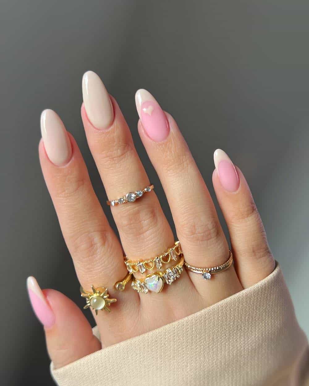 A hand with long almond nails painted with a soft beige featuring solid-colored nails, French tips, and heart details