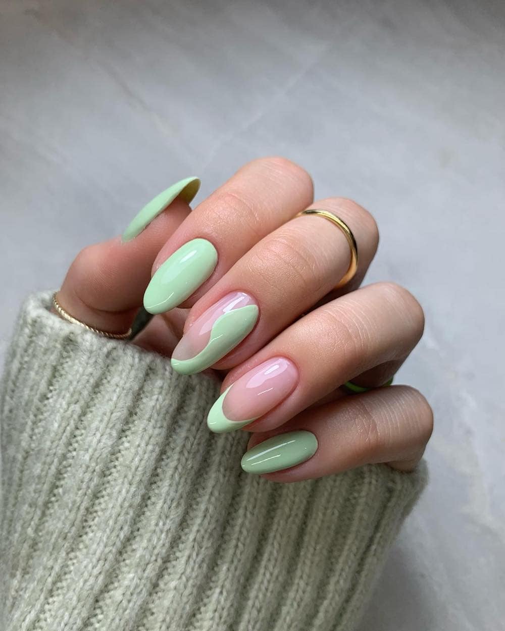A hand with long almond nails painted with a light spring green polish featuring solid-colored nails, French tips, and a wave accent nail