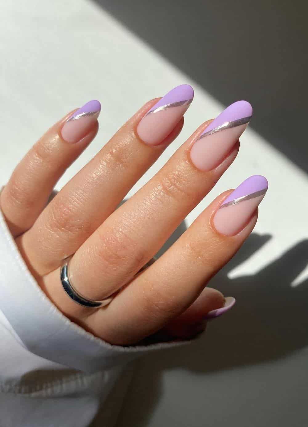 A hand with long nude almond nails with lavender side French tips with silver borders