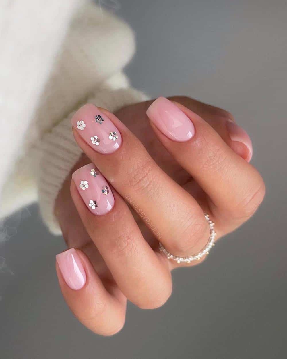 A hand with short nude square nails with two accent nails featuring silver flowers