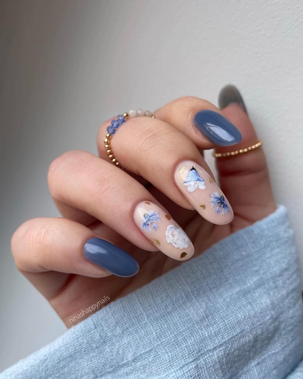 A hand with medium round nails painted a moody blue with nude accent nails featuring blue floral art