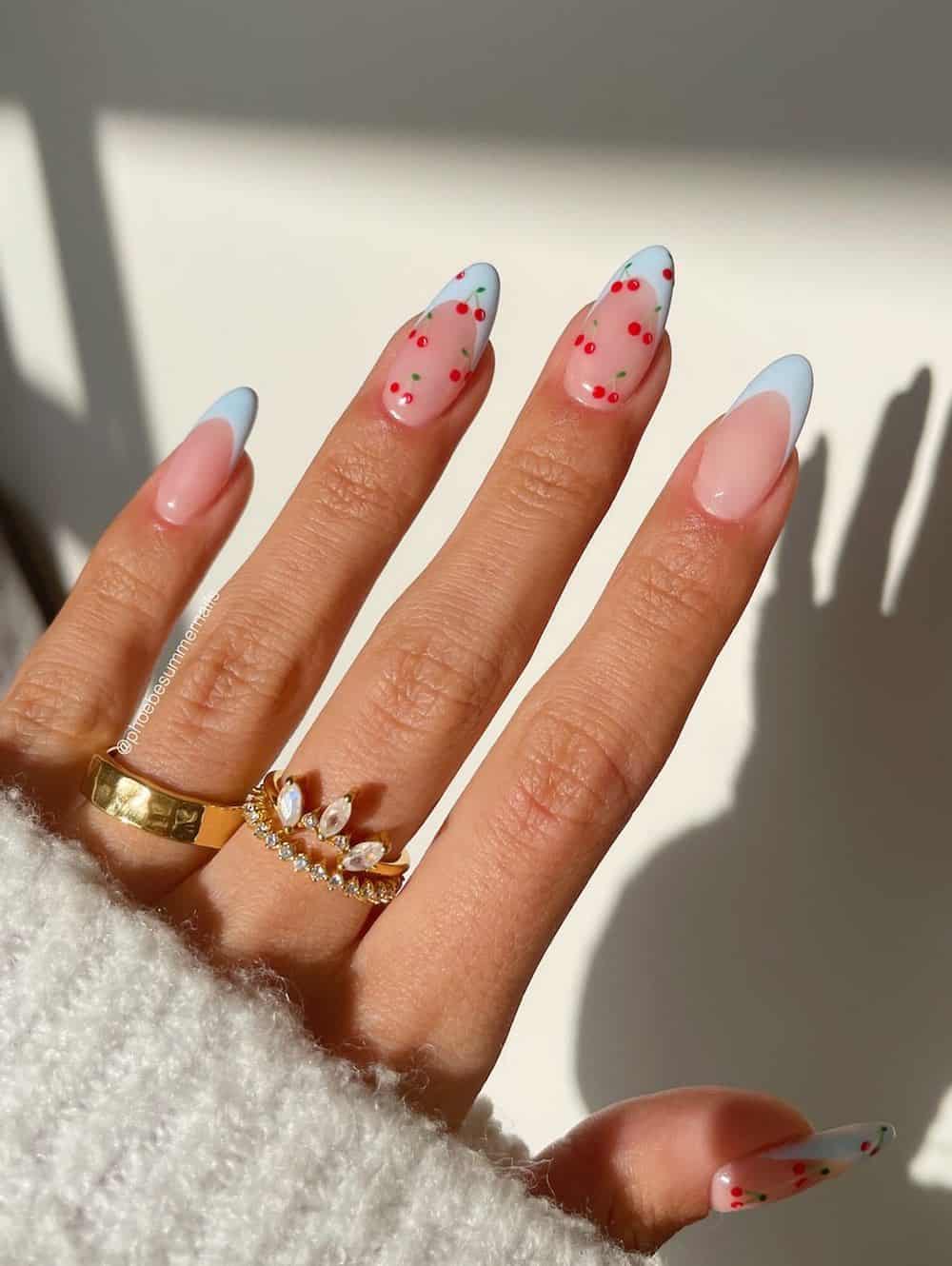 A hand with long nude almond nails with light blue French tips and cherry nail art on two nails
