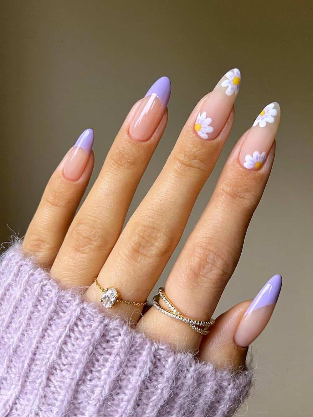 A hand with long nude almond nails with lilac French tips and two accent nails featuring white and purple floral art