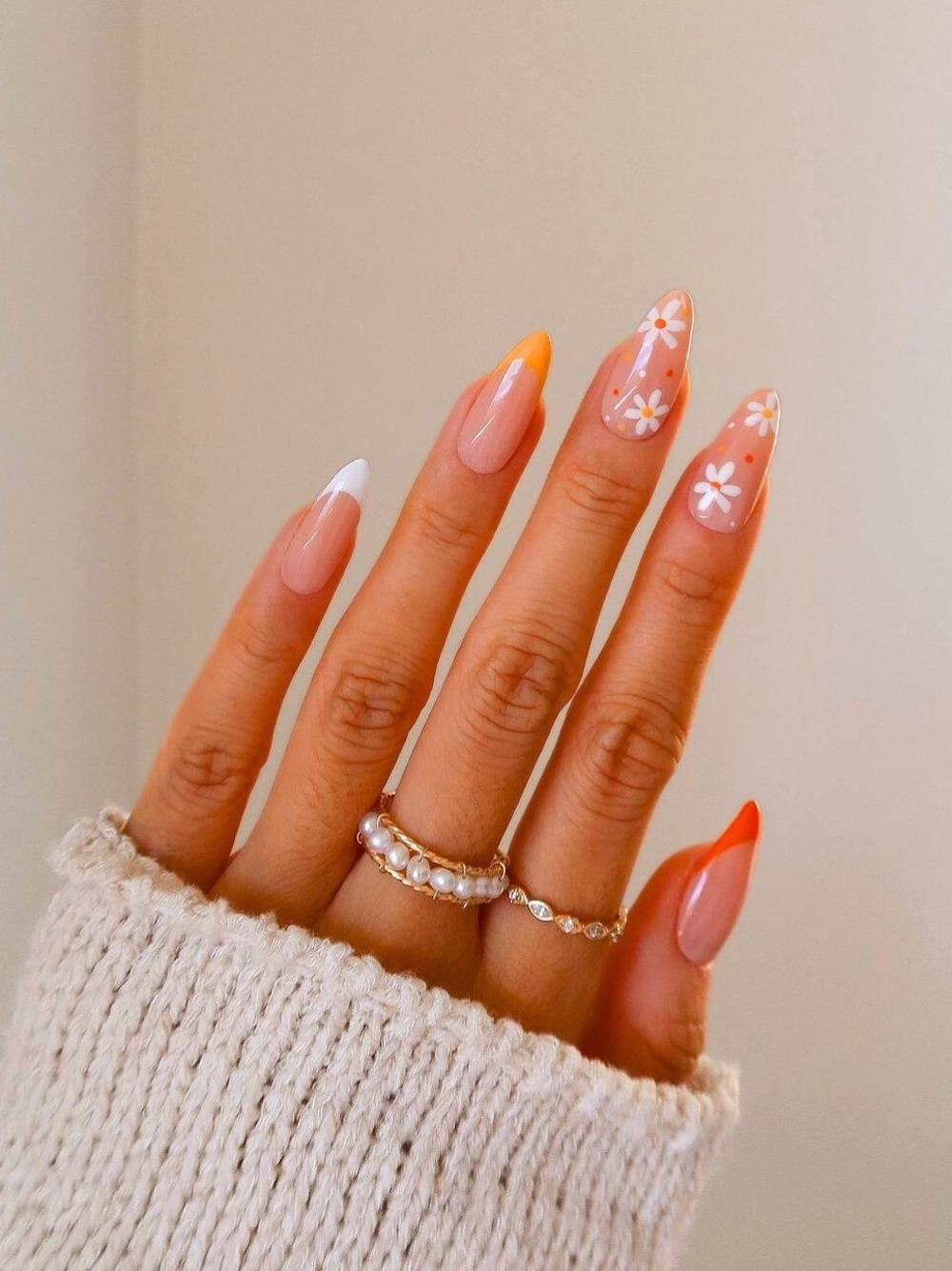 A hand with long nude almond nails featuring white, yellow, and orange French tips and accent nails with white floral art