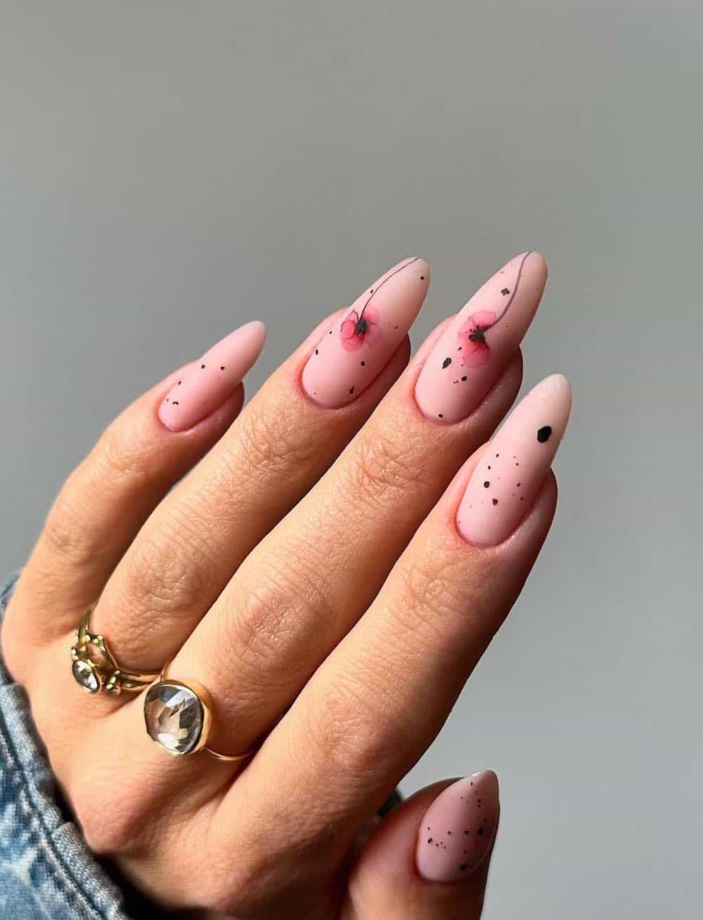A hand with long nude matte almond nails featuring black speckles and pink nail art