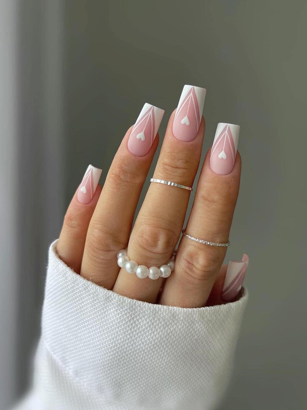 A hand with medium square nails painted a matte pink with white pointed French tips, white outlines and hearts
