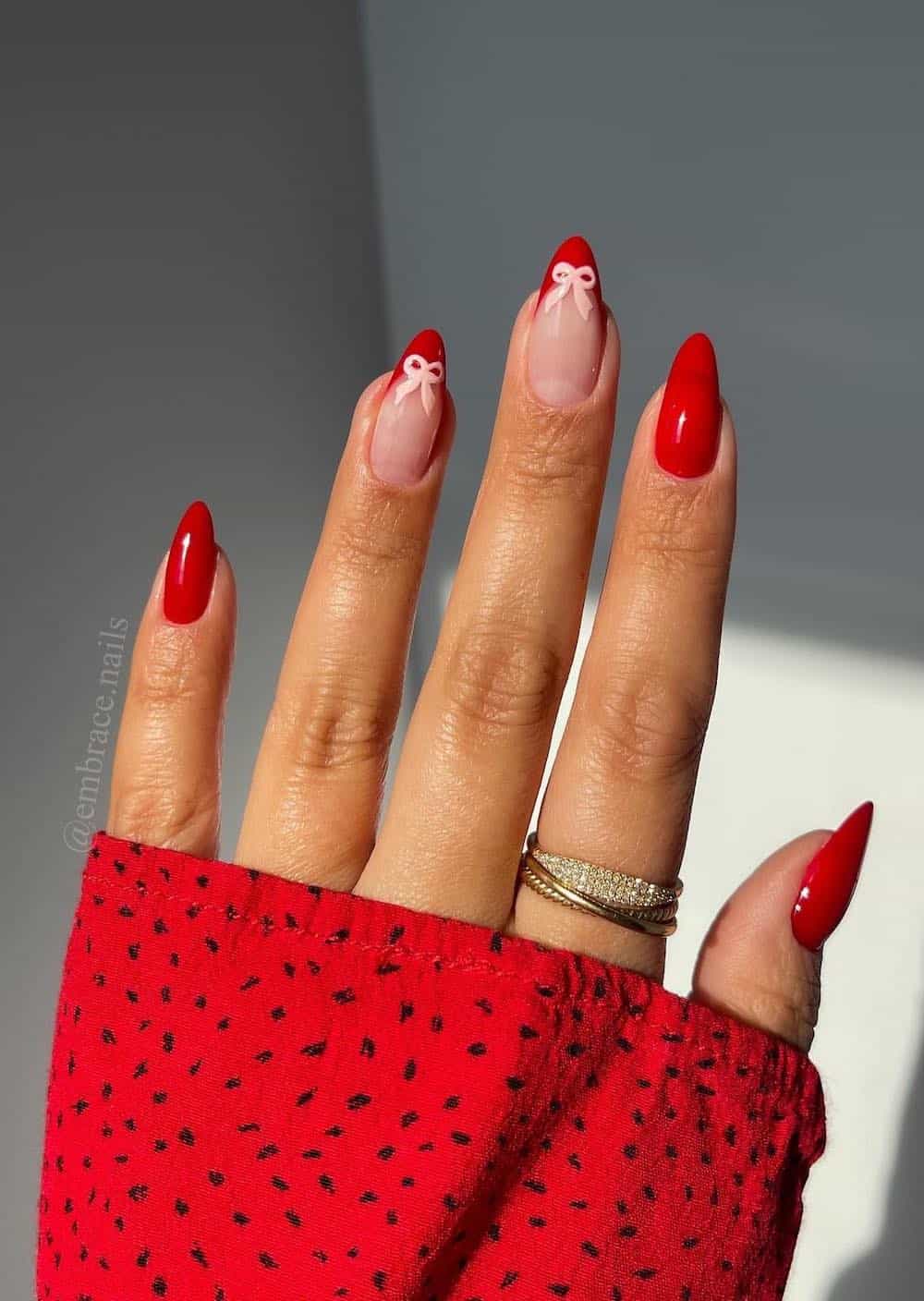 A hand with long almond nails painted a bright red with two red French tipped accents with pink bows