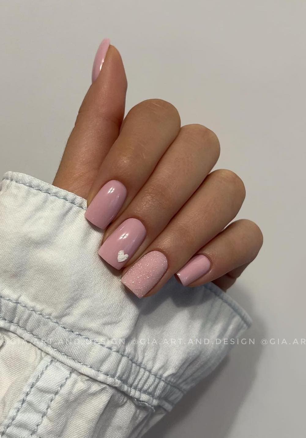 A hand with short square nails painted a dusty pink with a glitter coated accent nail and a white heart detail