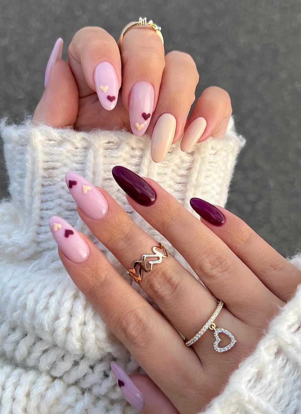 A hand with long almond nails painted a nude pink with beige and deep red accent nails and heart details