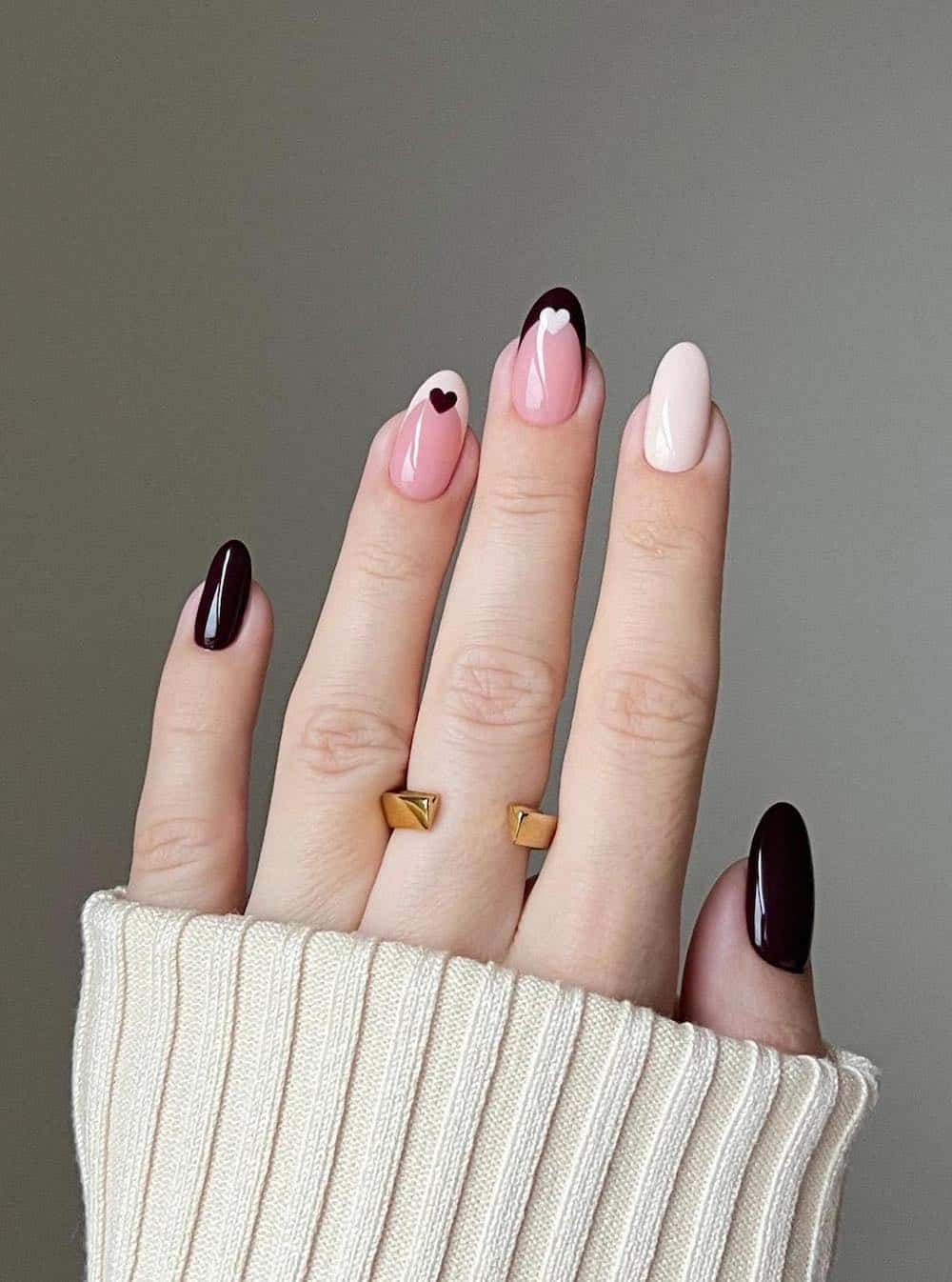 A hand with short round nails painted in cream, black, and nude pink accent nails with contrasting French tips and hearts