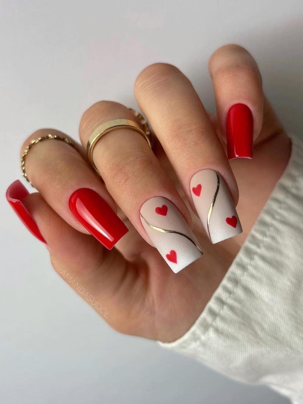 A hand with long square nails painted a bright red with two matte cream accent nails featuring gold waves and red hearts
