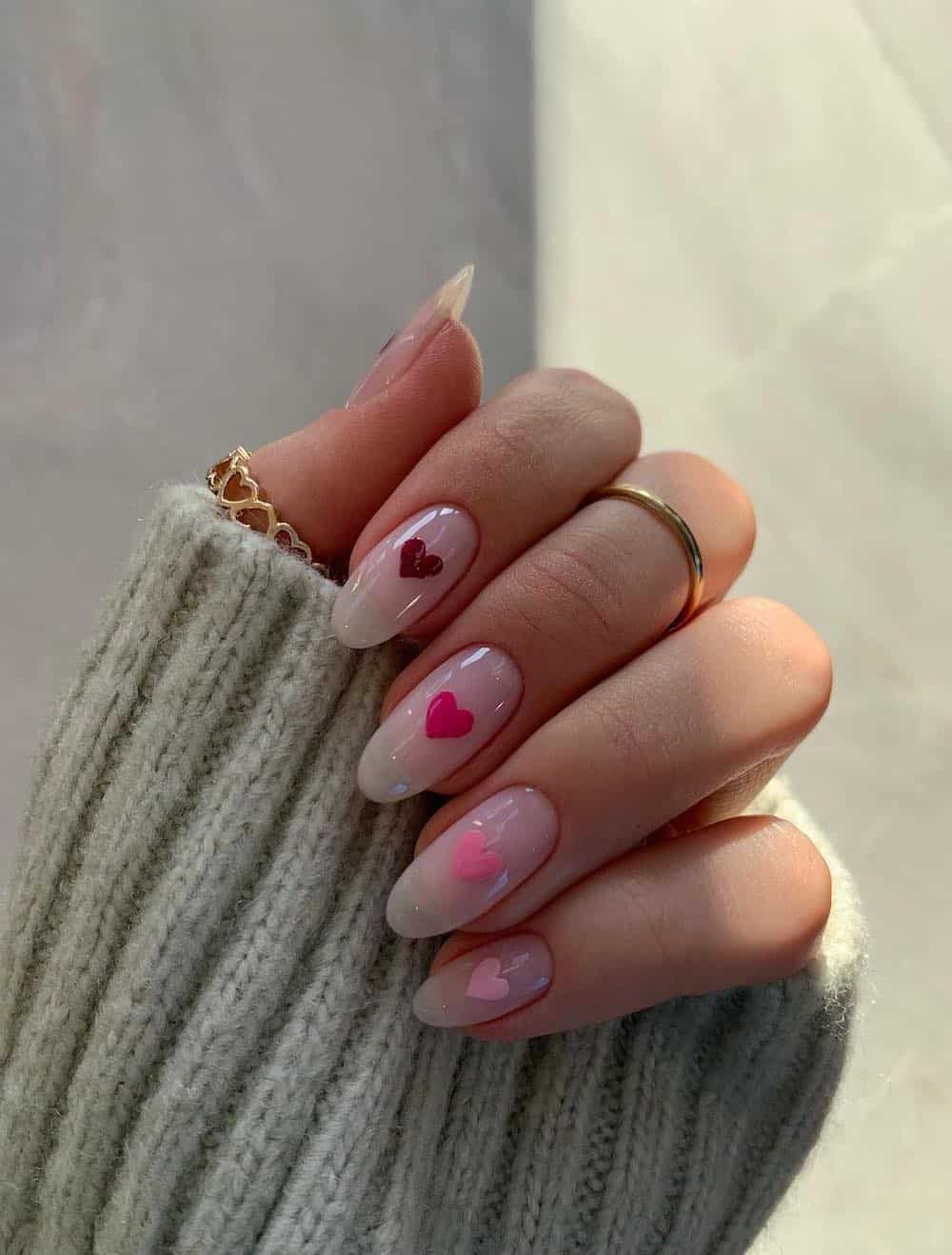 A hand with medium nude almond nails featuring gradient pink and red hearts