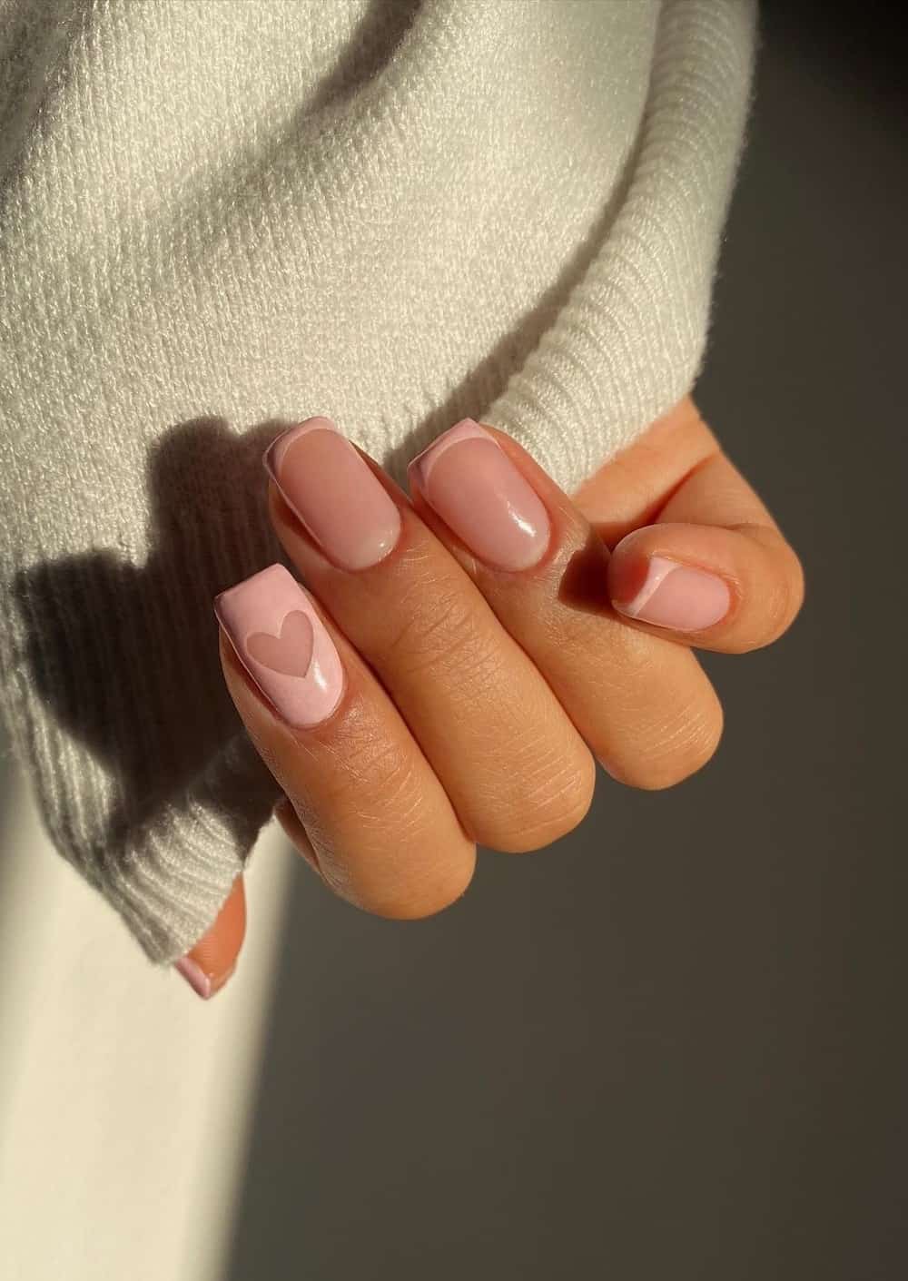 A hand with short square nails painted with light pink French tips and a light pink accent nail with a negative space heart