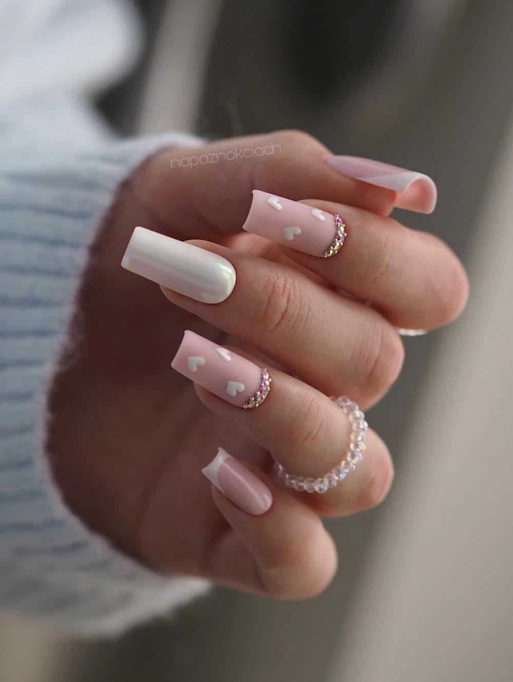A hand with long square nails featuring a chrome white nail, light pink nails with white tips, and matte light pink nails with white hearts and gem accents