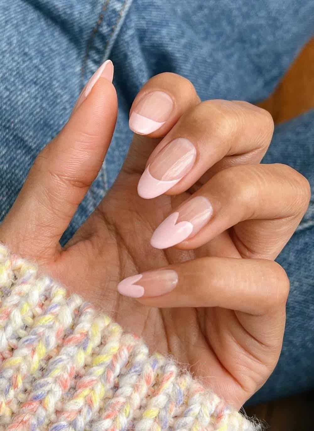 A hand with long almond nails painted with pastel pink French tips and two accent nails with heart shaped French tips