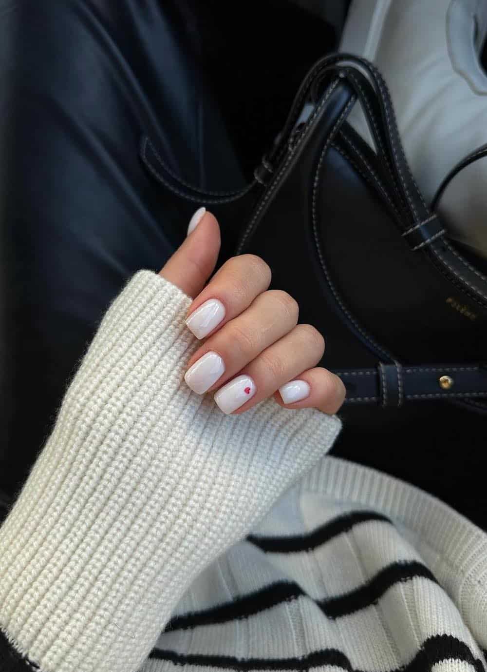 A hand with short square white nails with a red heart on one accent nail