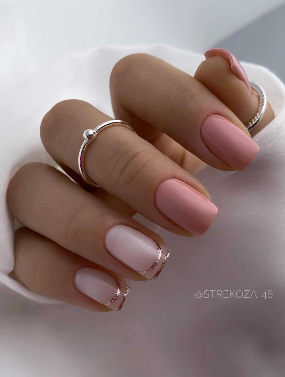 A hand with short square nails painted a dusty pink with two milky white accent nails featuring glittering rose gold French tip outlines