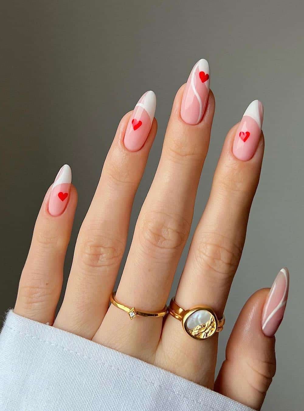 A hand with long nude pink almond nails with wavy white French tips and red hearts