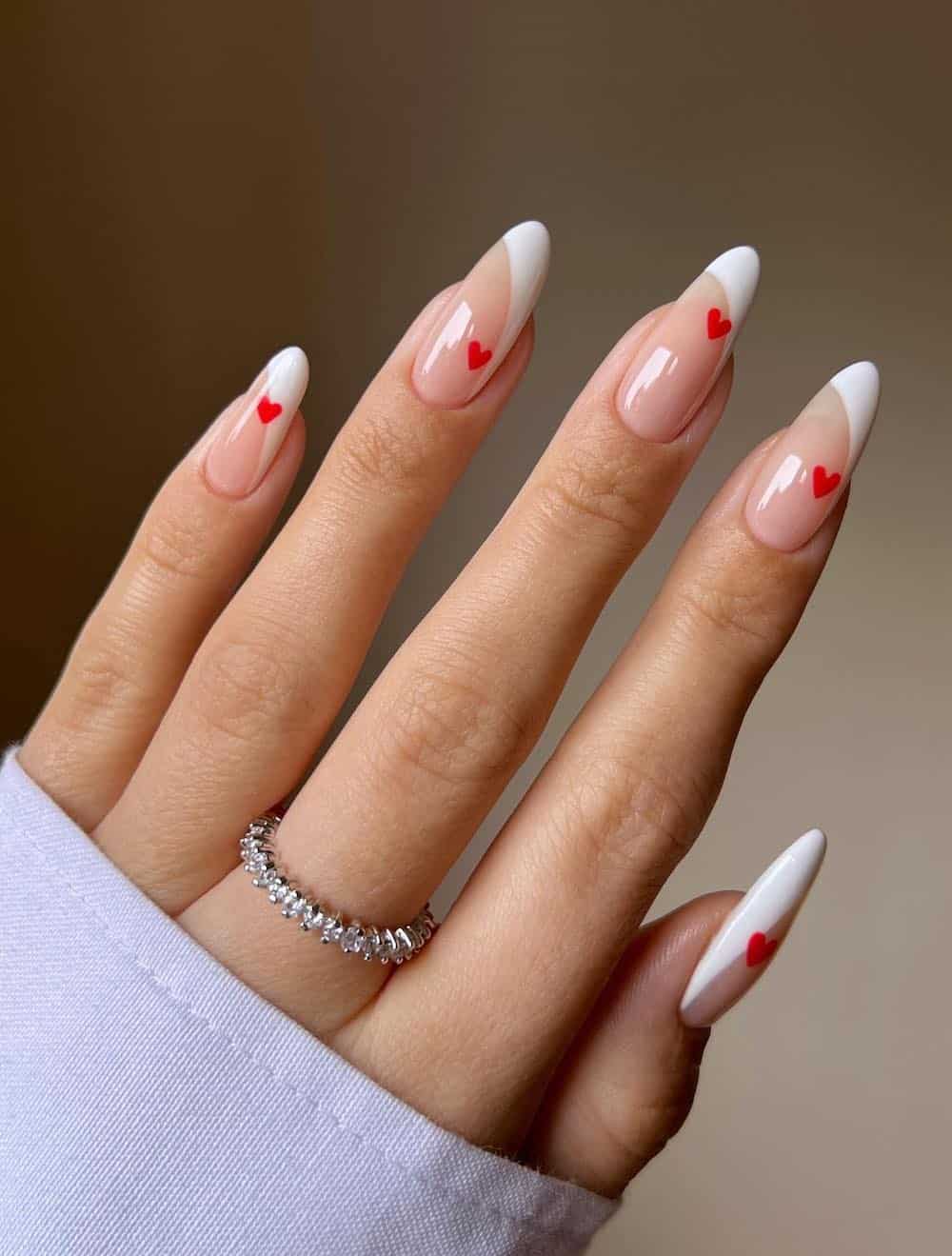 A hand with long almond nails painted a glossy nude with curved French tips and red hearts