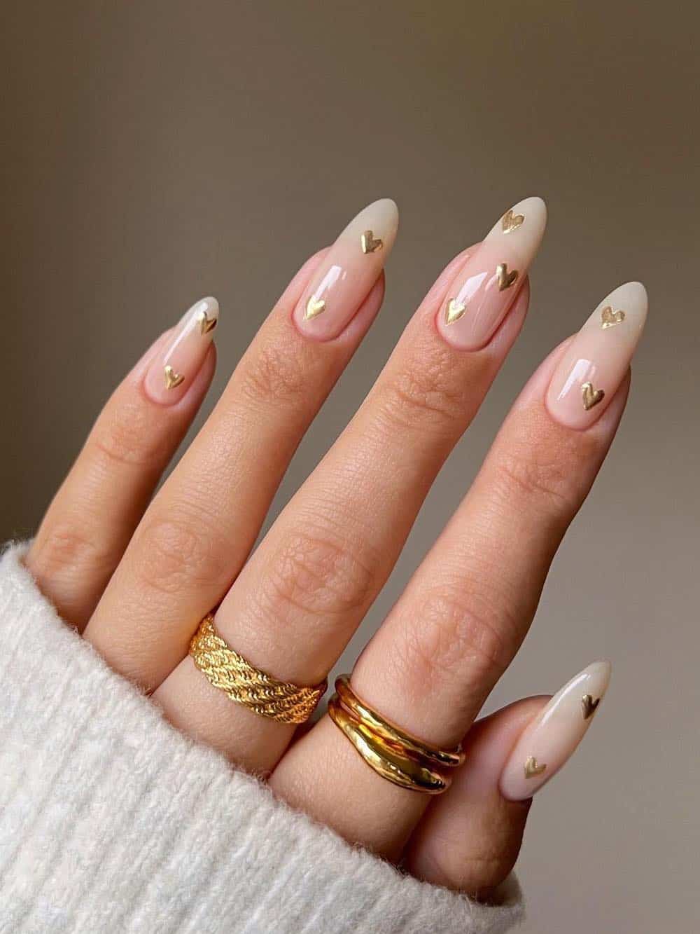 A hand with long nude almond nails with gold hearts