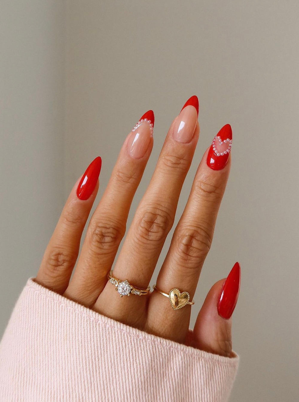 A hand with long nude almond nails with cherry red solid-colored nails and French tips with heart accents and pearl details