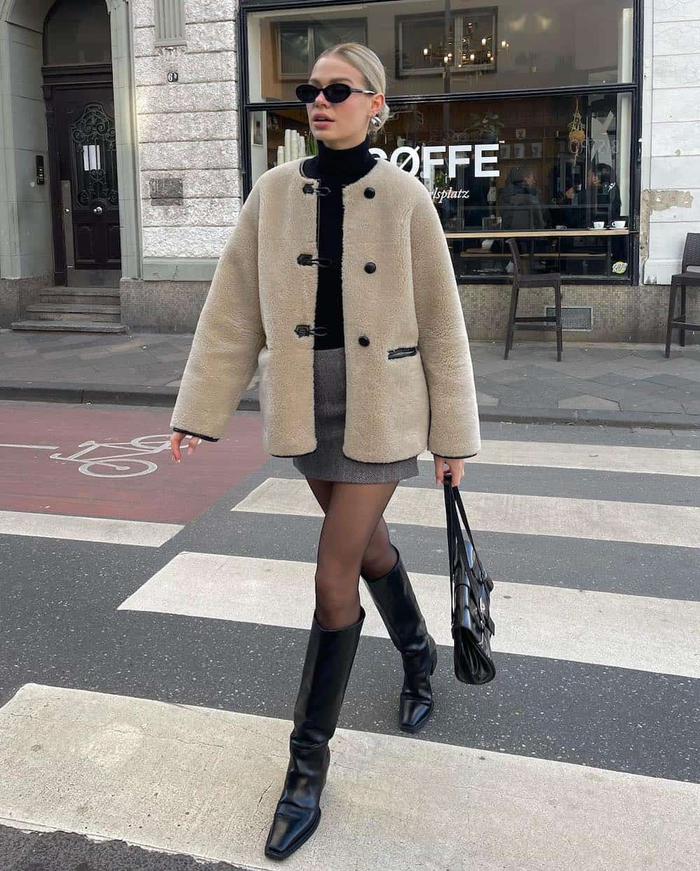 Woman wearing tall black boots, black tights, a grey mini skirt and a black turtleneck with a cream colored sherpa coat.