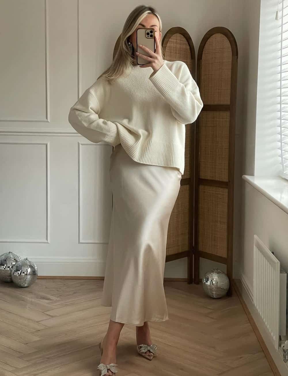 Woman wearing a cream colored midi slip skirt with a white sweater and sparkly bow stilettos.