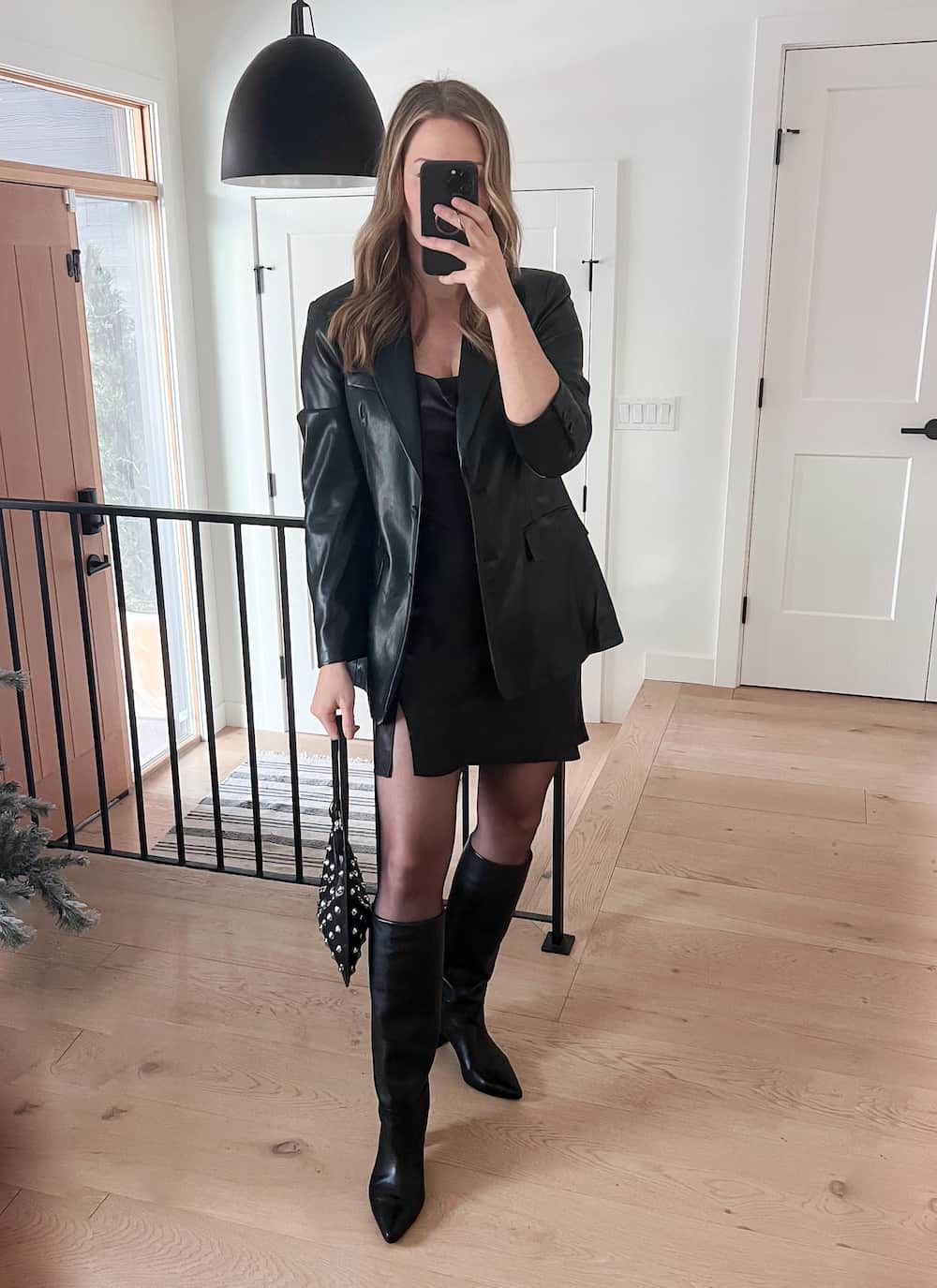 woman wearing a winter going out outfit with a black leather blazer over a black mini slip dress with black tights and knee-high boots