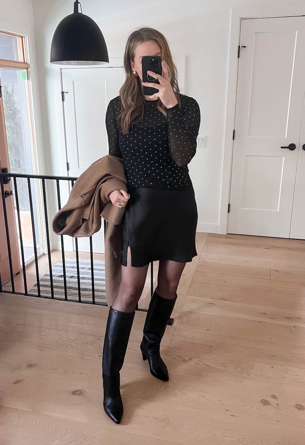 woman wearing a winter going out outfit with a long wool camel coat, black rhinestone long sleeve top, black mini skirt, black tights, and knee-high boots