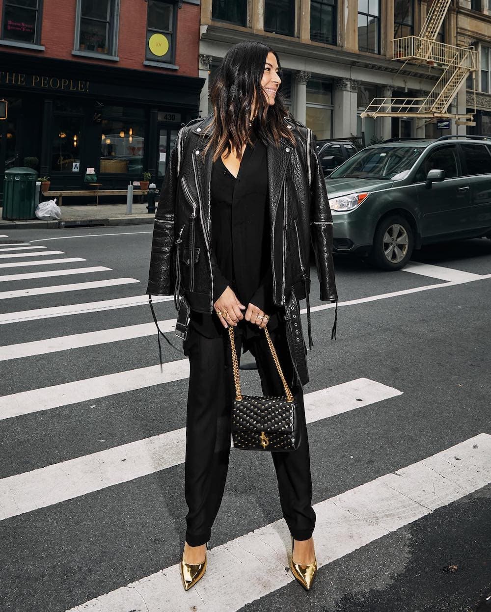 woman wearing a winter going out outfit with a black leather jacket over a black jumpsuit with gold pumps