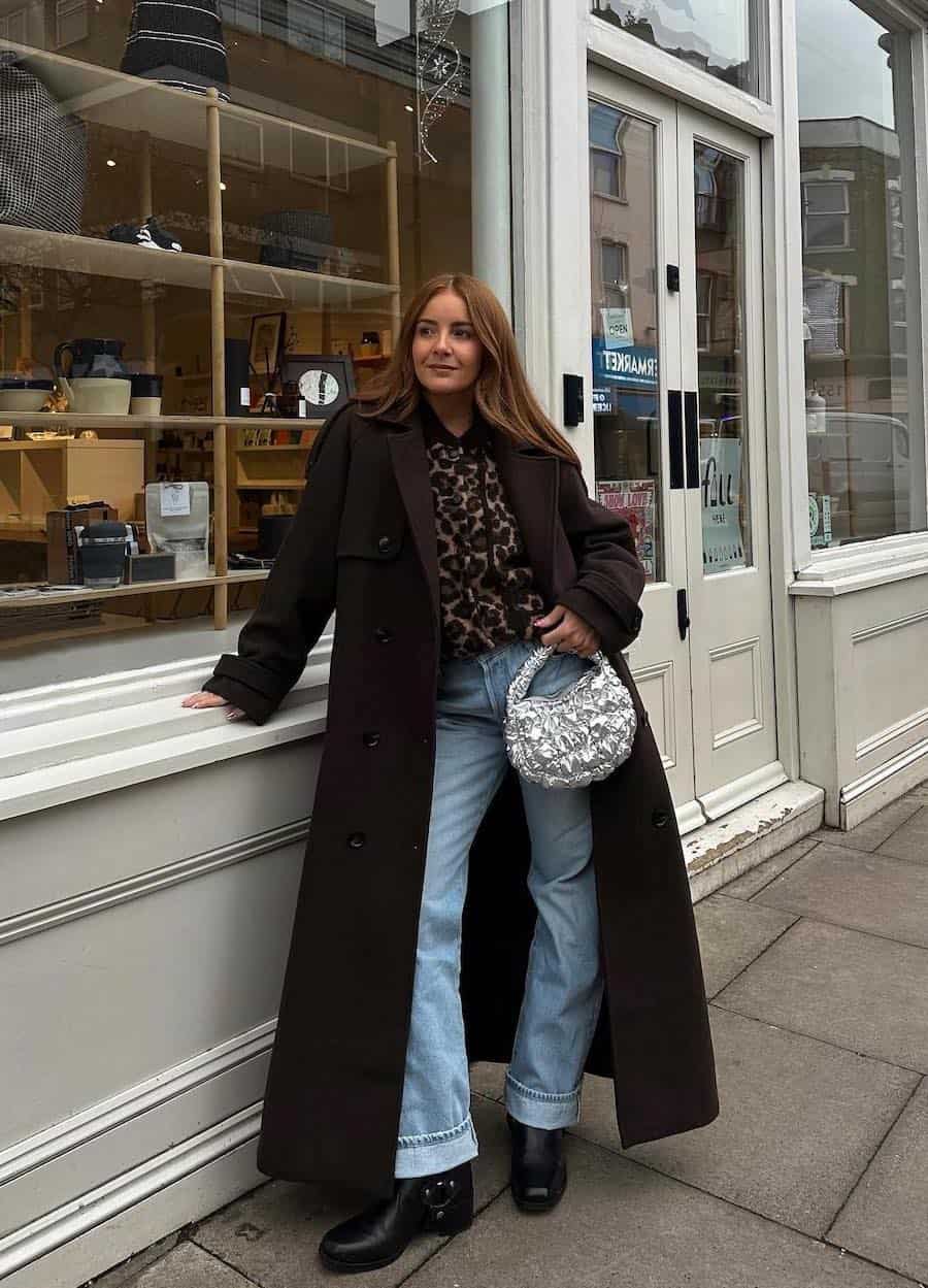 woman wearing a winter going out outfit with a long wool coat, leopard print blouse, blue jeans, and metallic silver bag 