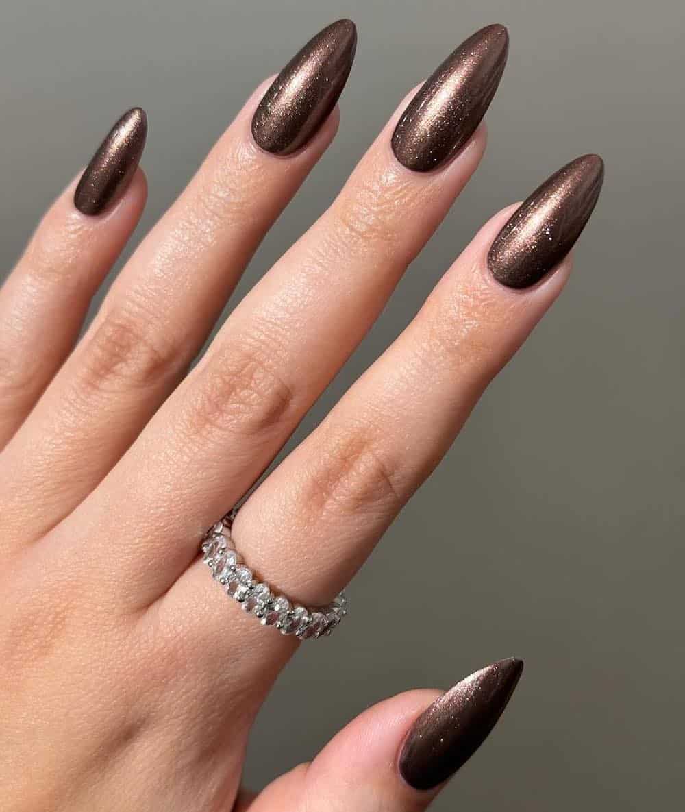 A hand with long almond nails painted a rich chocolate brown with a chrome finish