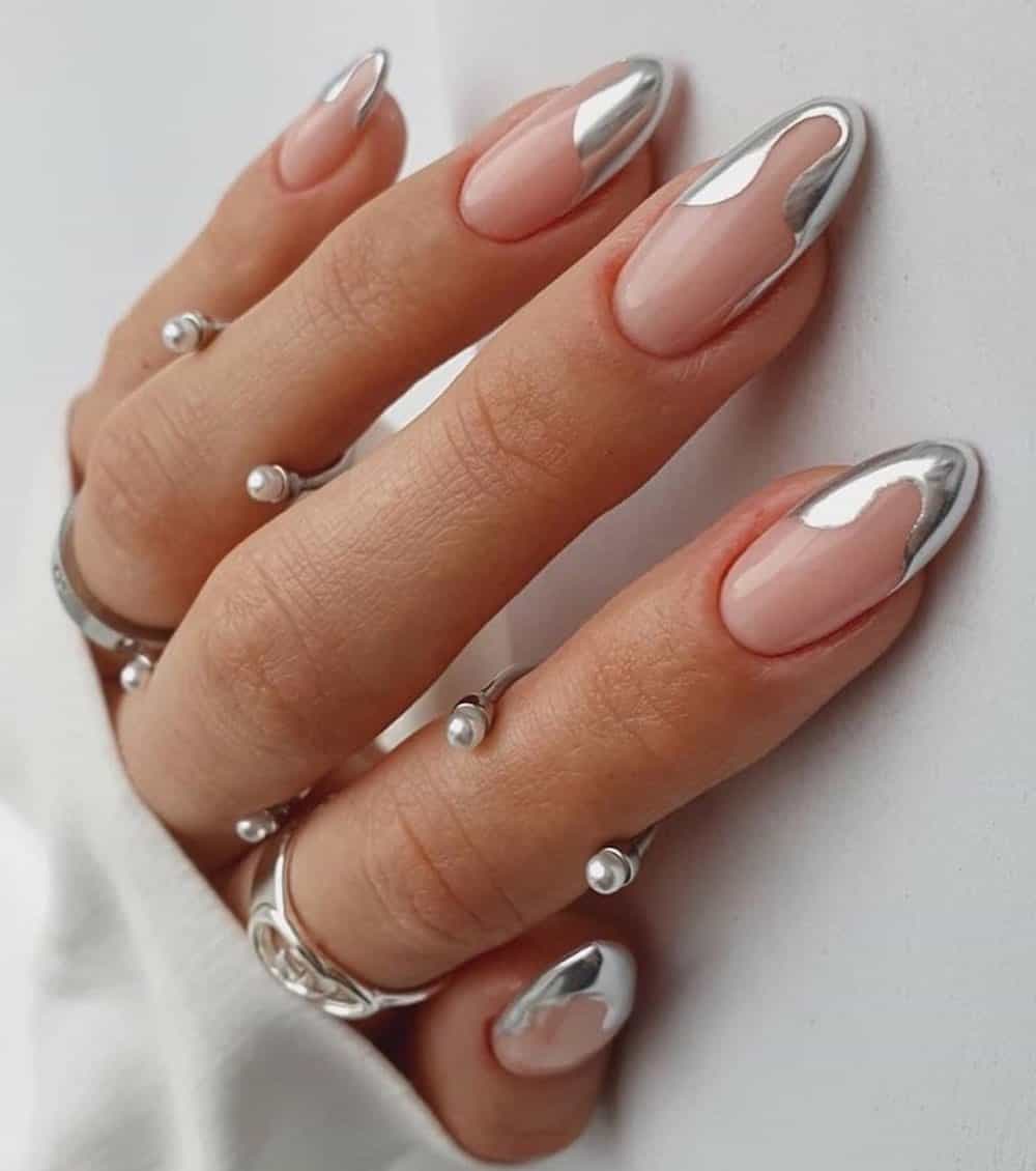 A hand with medium almond nails painted a glossy nude with wavy silver chrome French tips