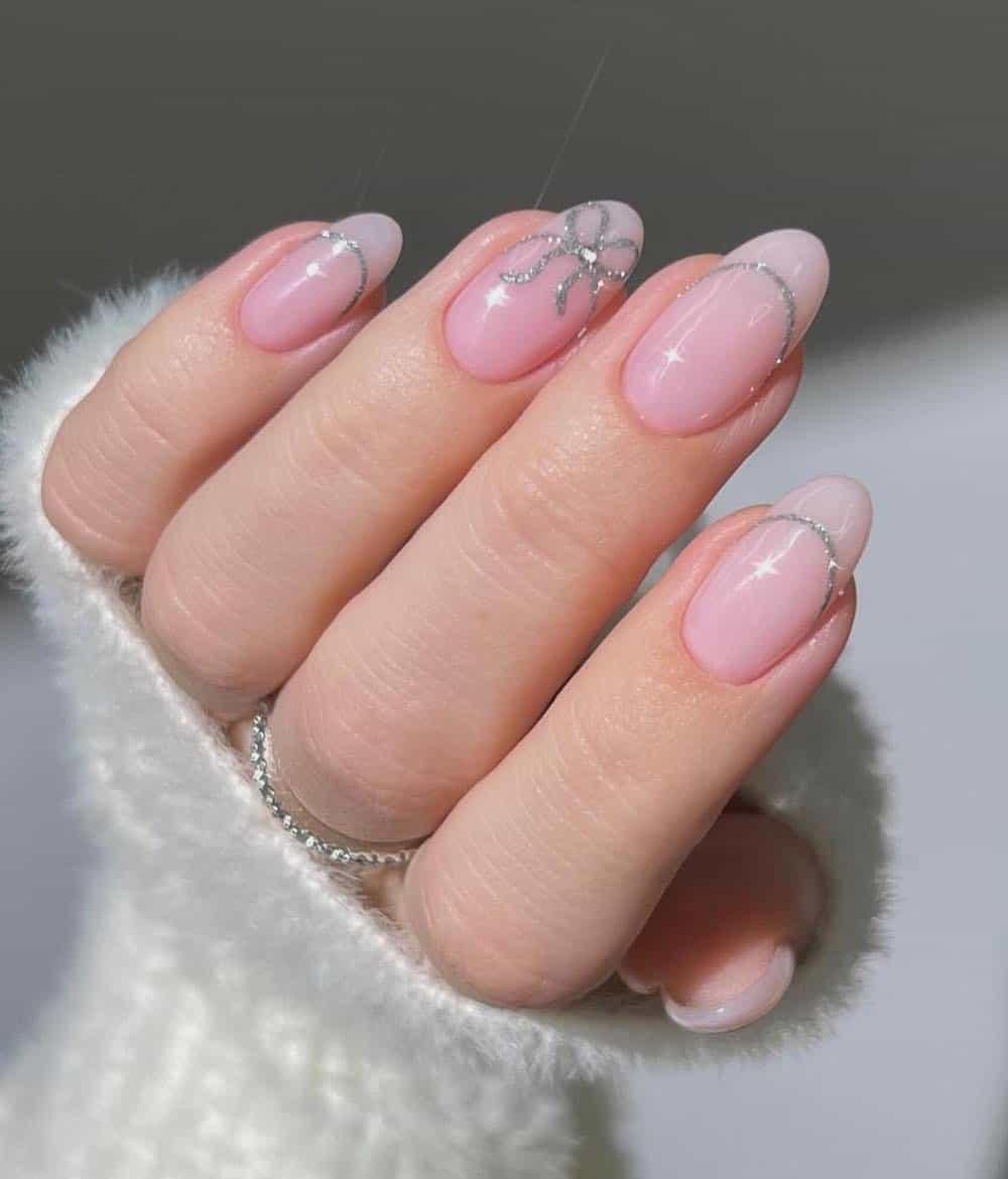 A hand with nude pink almond nails with a sparkling silver French tip outlines and a bow detail