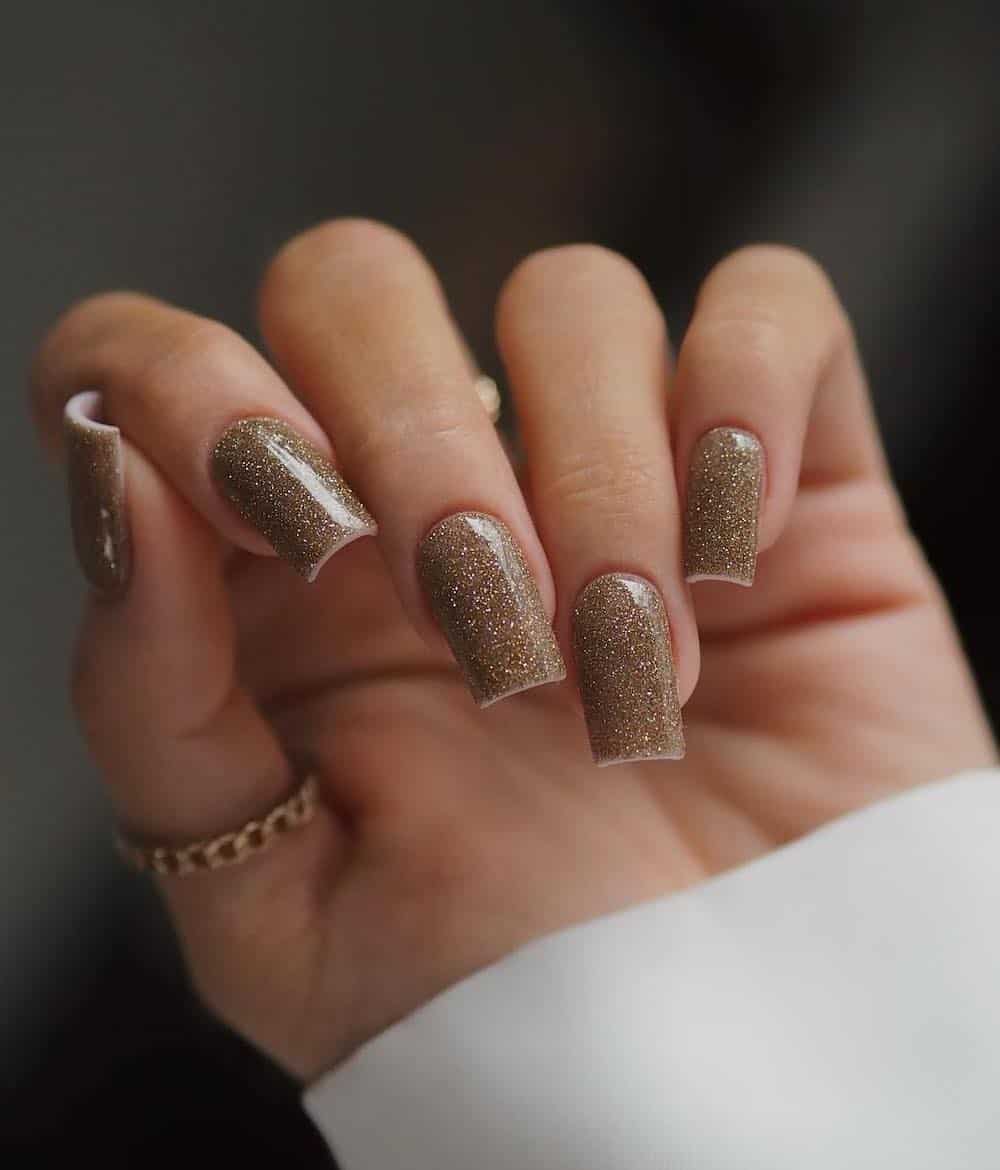 A hand with long square nails painted with a glittering gold nail polish