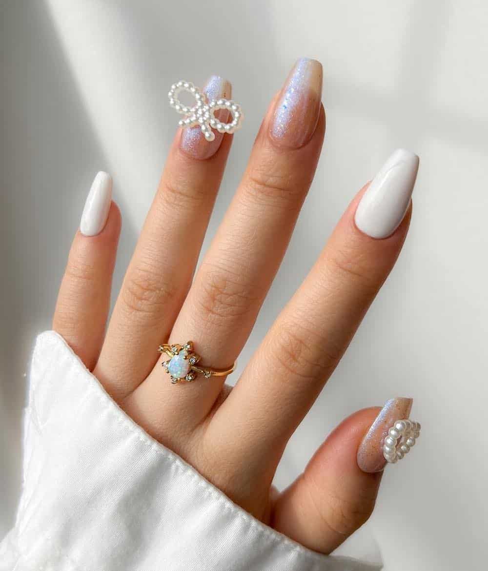 A hand with medium coffin nails painted white and glittery iridescent polish with pearl bead accents in the shape of a bow and heart