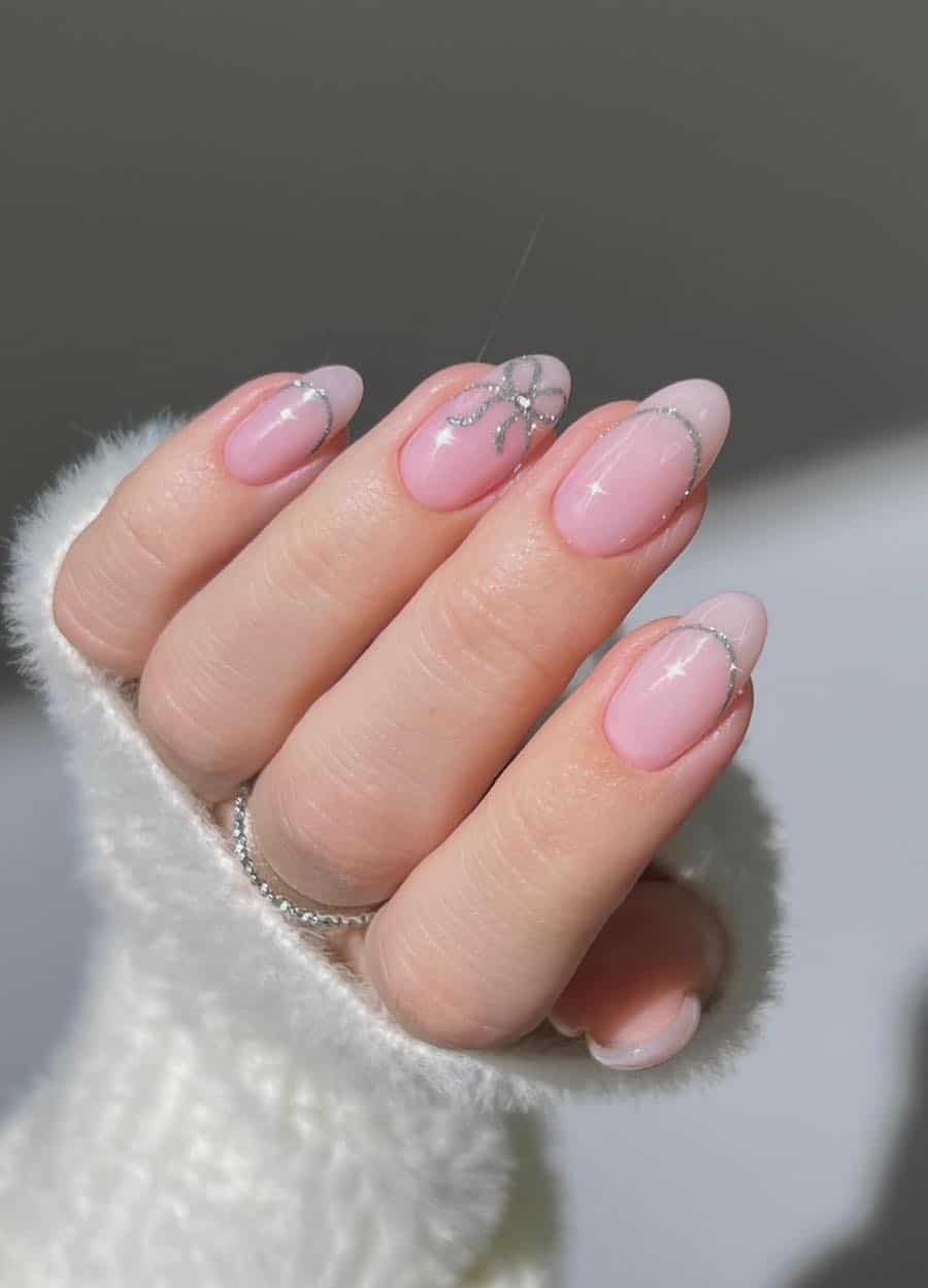 Medium round nails painted a glossy light pink with silver glitter French tip outlined and a bow accent nail