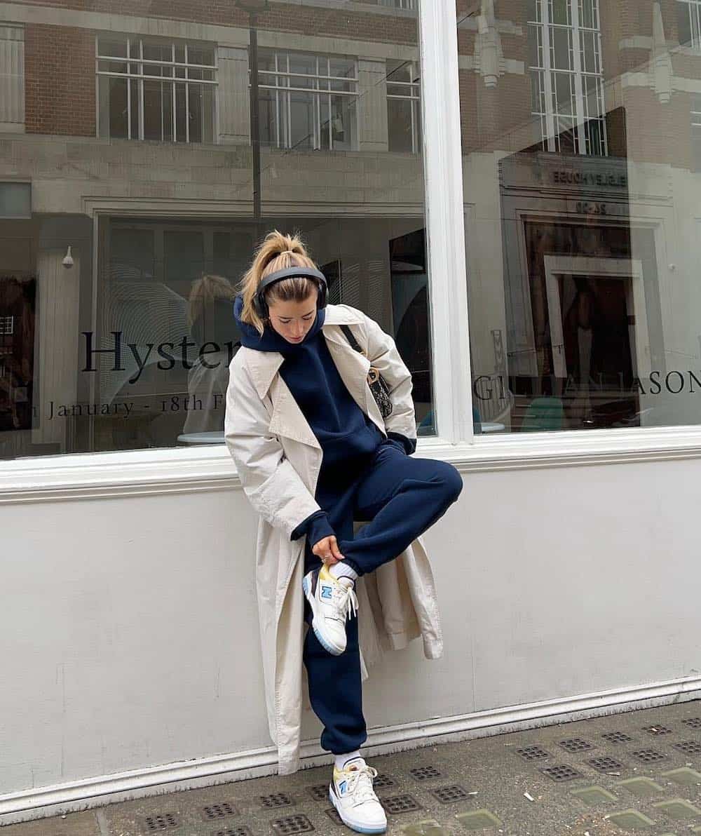 Woman wearing a navy sweatsuit set with a cream colored trench coat and white sneakers.