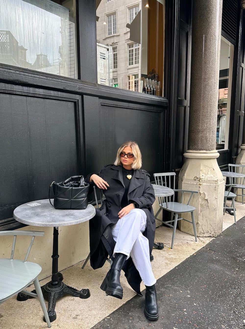 Woman sitting at a cafe wearing light wash jeans, lugg boots, a black knit top and a black trench coat.