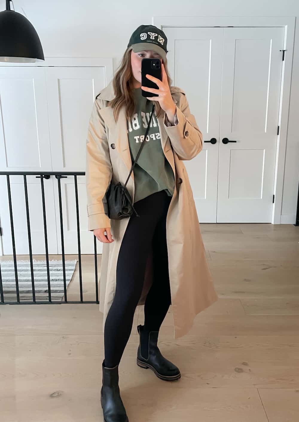 Christal wearing black leggings, a green sweatshirt, black Chelsea boots and a tan trench coat with a baseball cap.