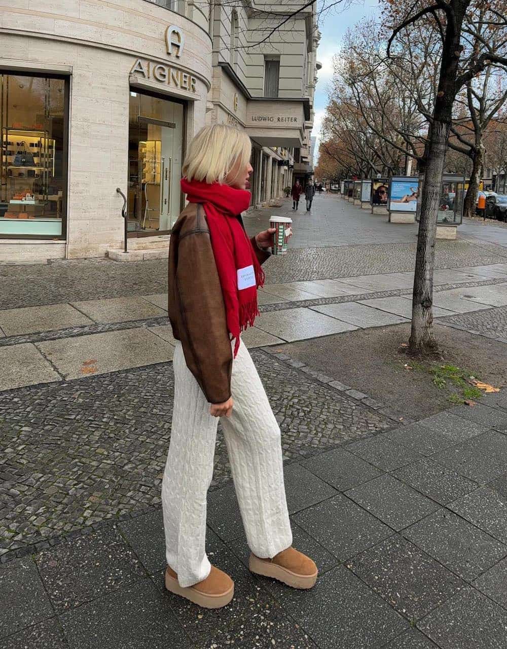 Woman wearing white knit pants, platform Ugg boots, a brown leather bomber jacket and a red scarf.